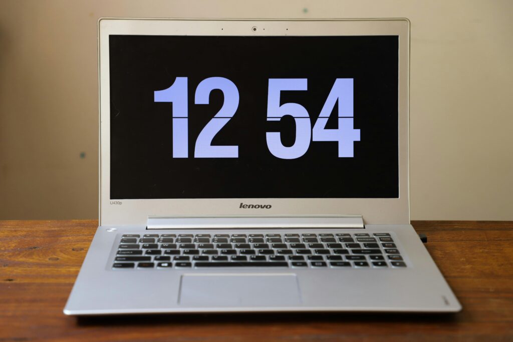 Silver laptop on a wooden desk showing a digital clock at 12:54.