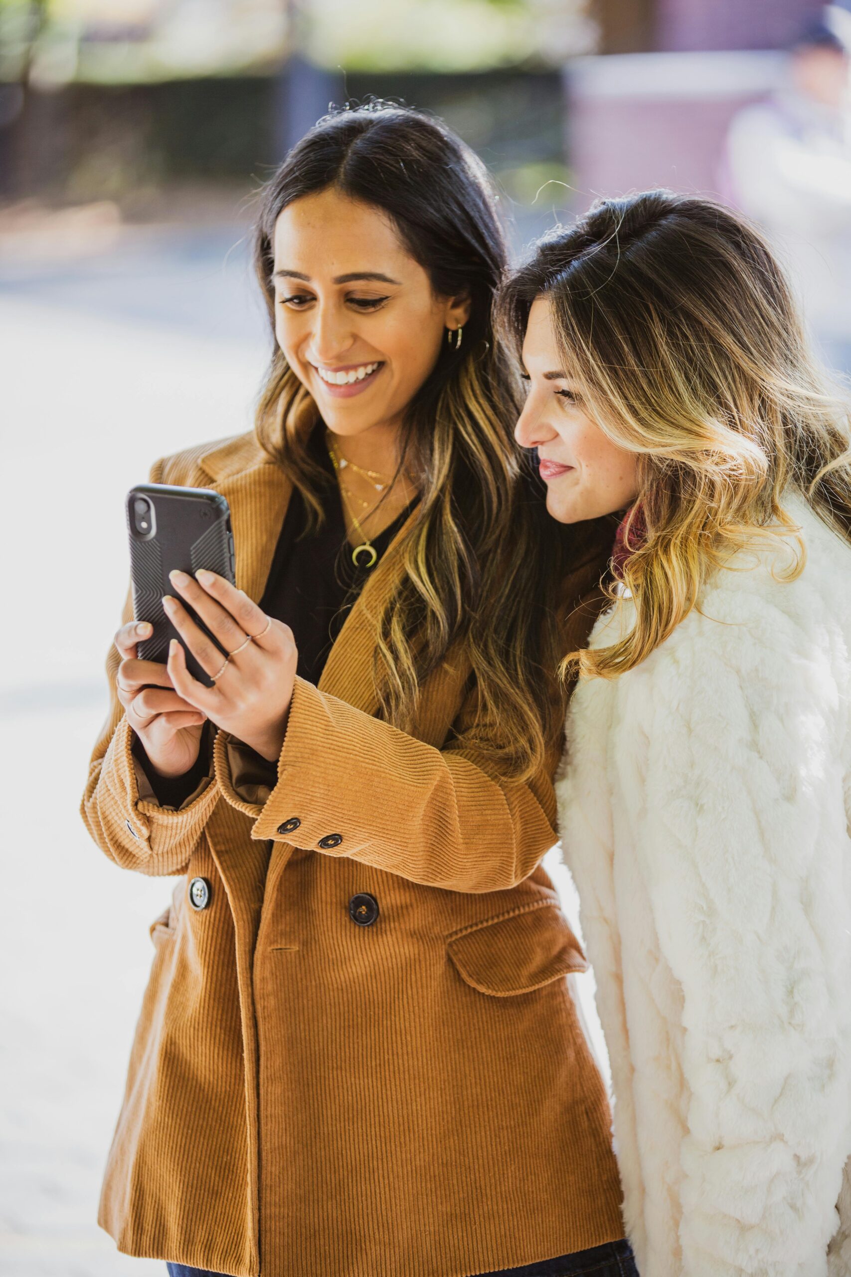 Two fashionable women smilingly share an enjoyable moment together while using a smartphone outdoors.