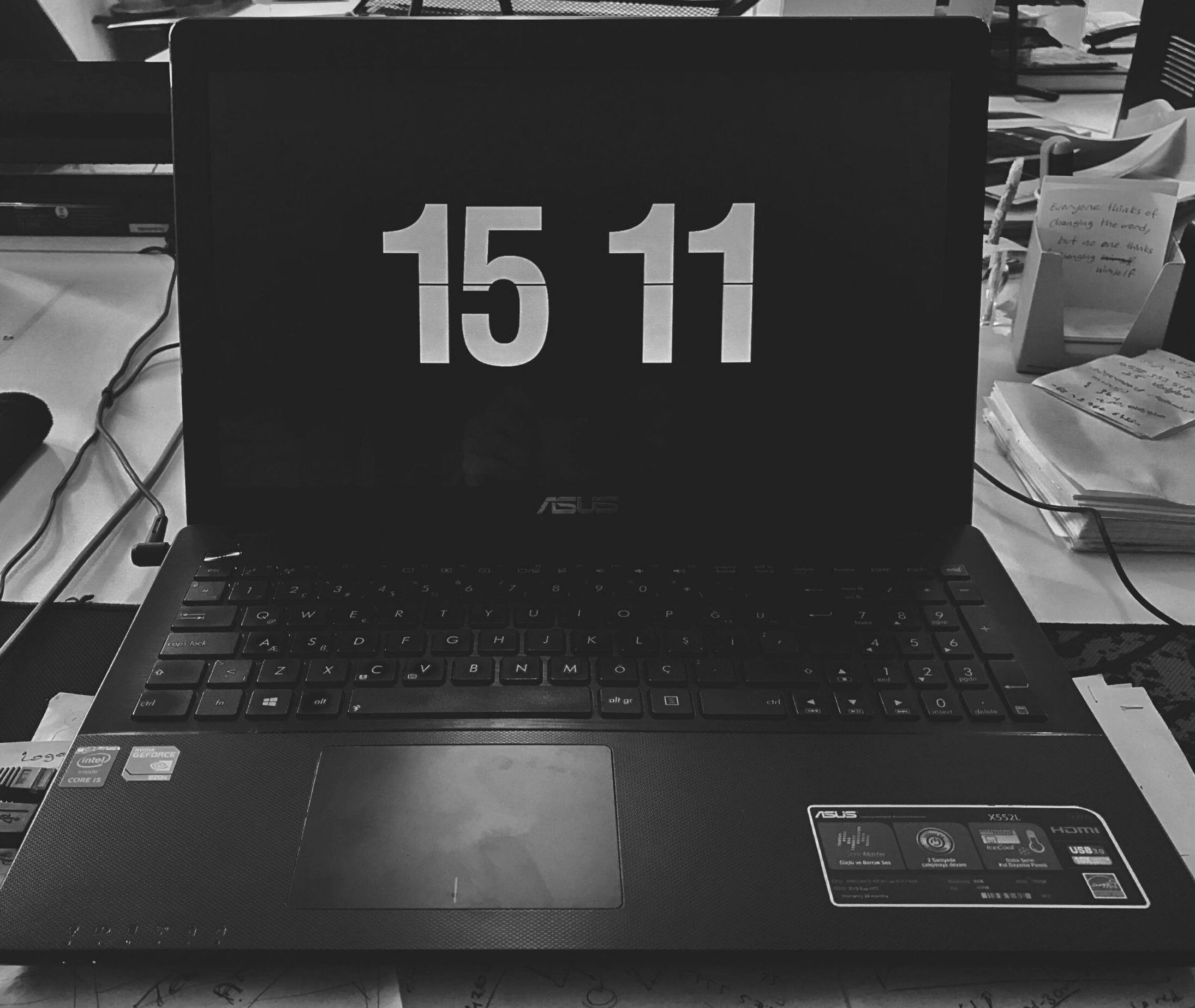 Black and white photo of a laptop displaying time, surrounded by desk items in a workspace.