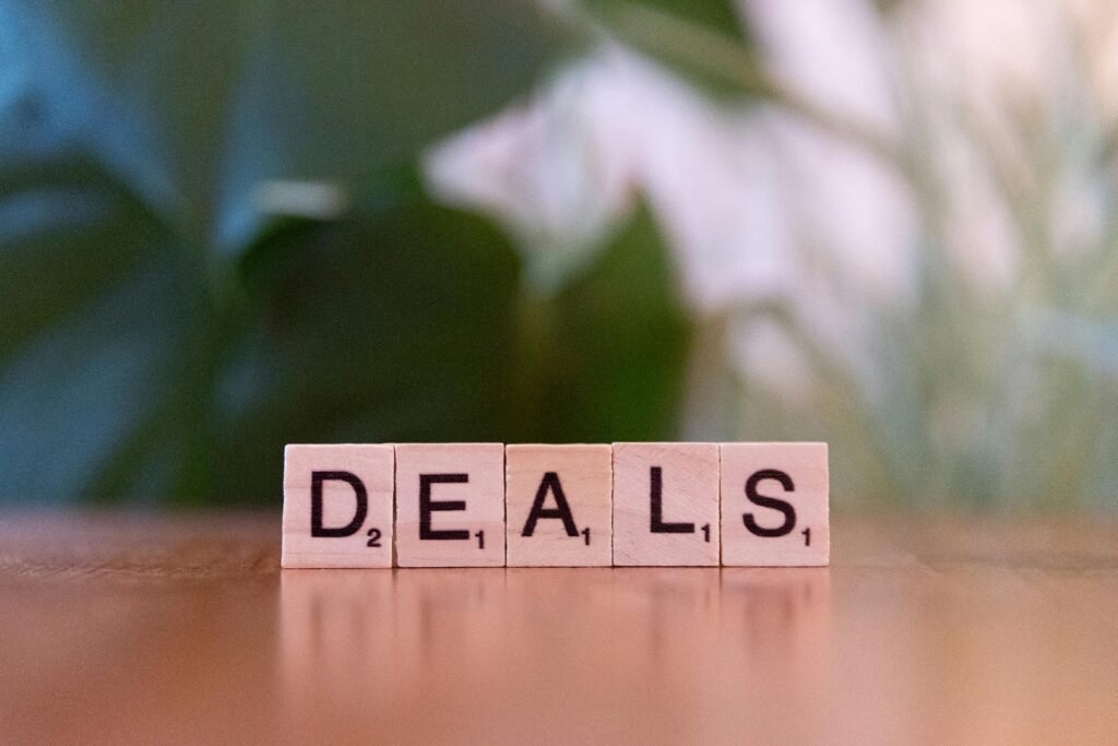 Close-up of Scrabble tiles spelling 'DEALS' on a wooden surface with blurry green background.